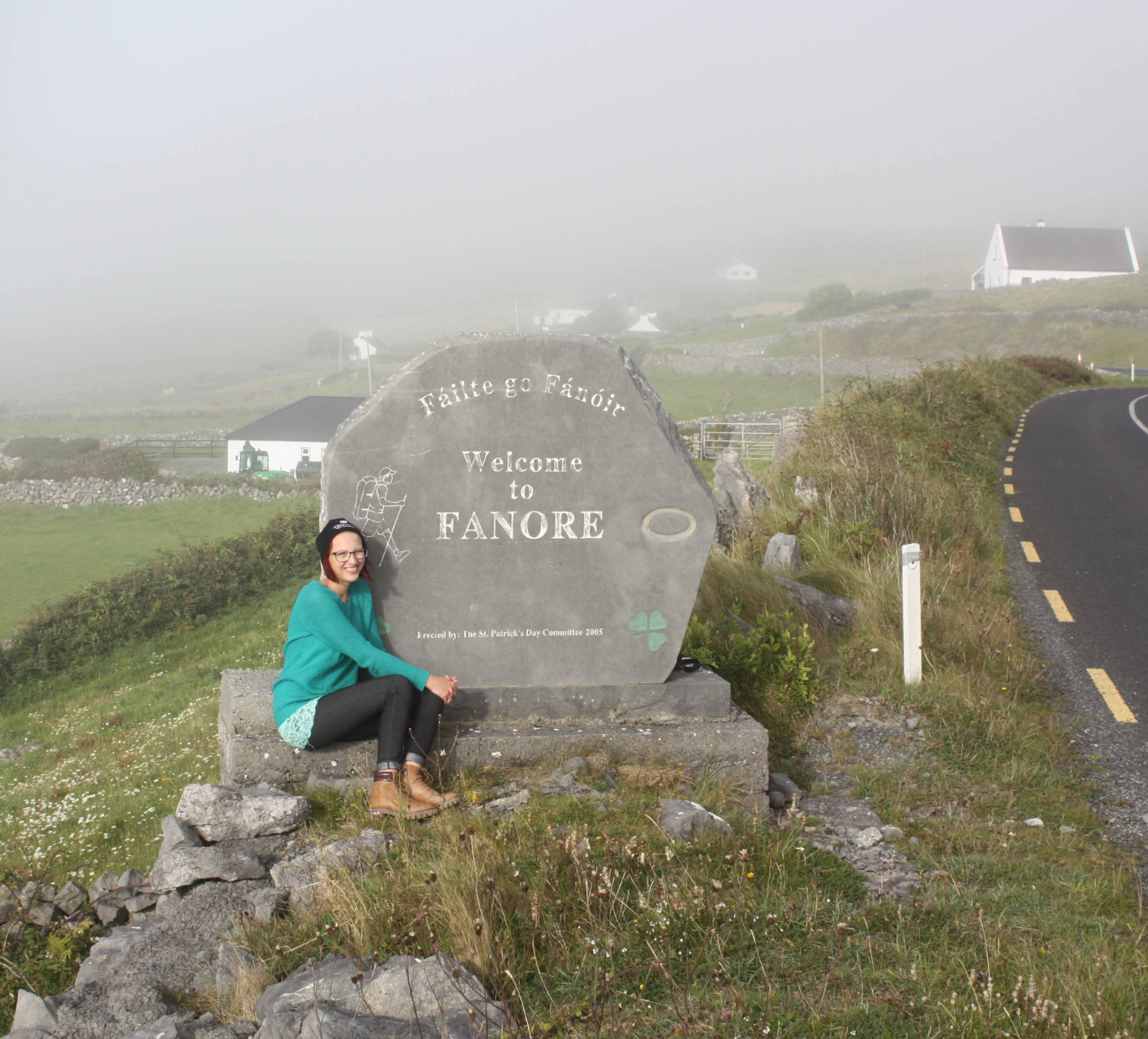 Autorin Jo Jonson sitzt lächelnd an einem Stein, der das Ortseingangsschild von Fanore in Westirland ist. Im HIntergrund sieht man einige Häuser im Nebel und eine Straße
