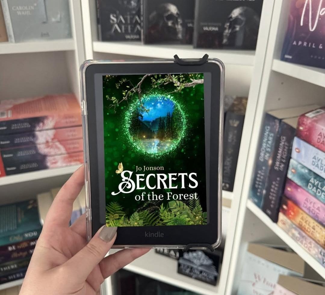 Die Romantasy-Neuerscheinung "Secrets of the Forest" von Jo Jonson wird als Ebook in die Kamera gehalten. Im Hintergrund sieht man ein Bücherregal