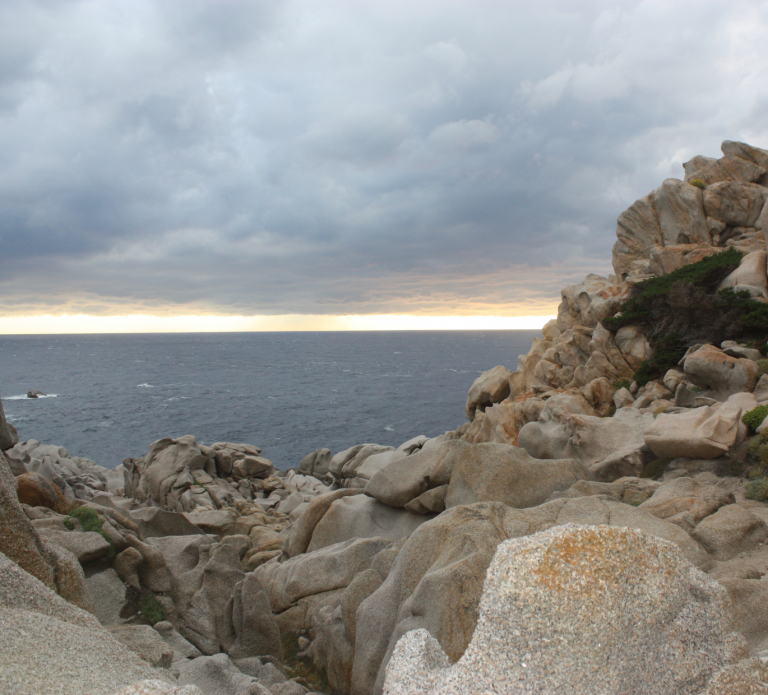Scharfe Felsen, dahinter das Meer und ein orangener Streifen am Horizont für den Sonnenuntergang im Urlaub auf Sardinien