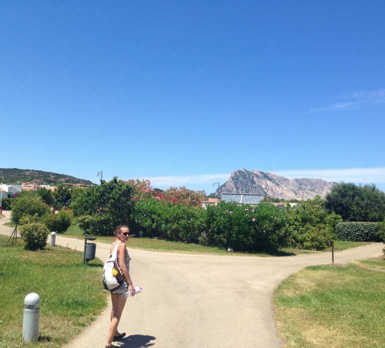 Jo Jonson steht auf einem Weg und dreht sich zum Fotografen um. Über ihr strahlend blauer Himmel und im Hintergrund ein Berg beim Urlaub auf Sardinien