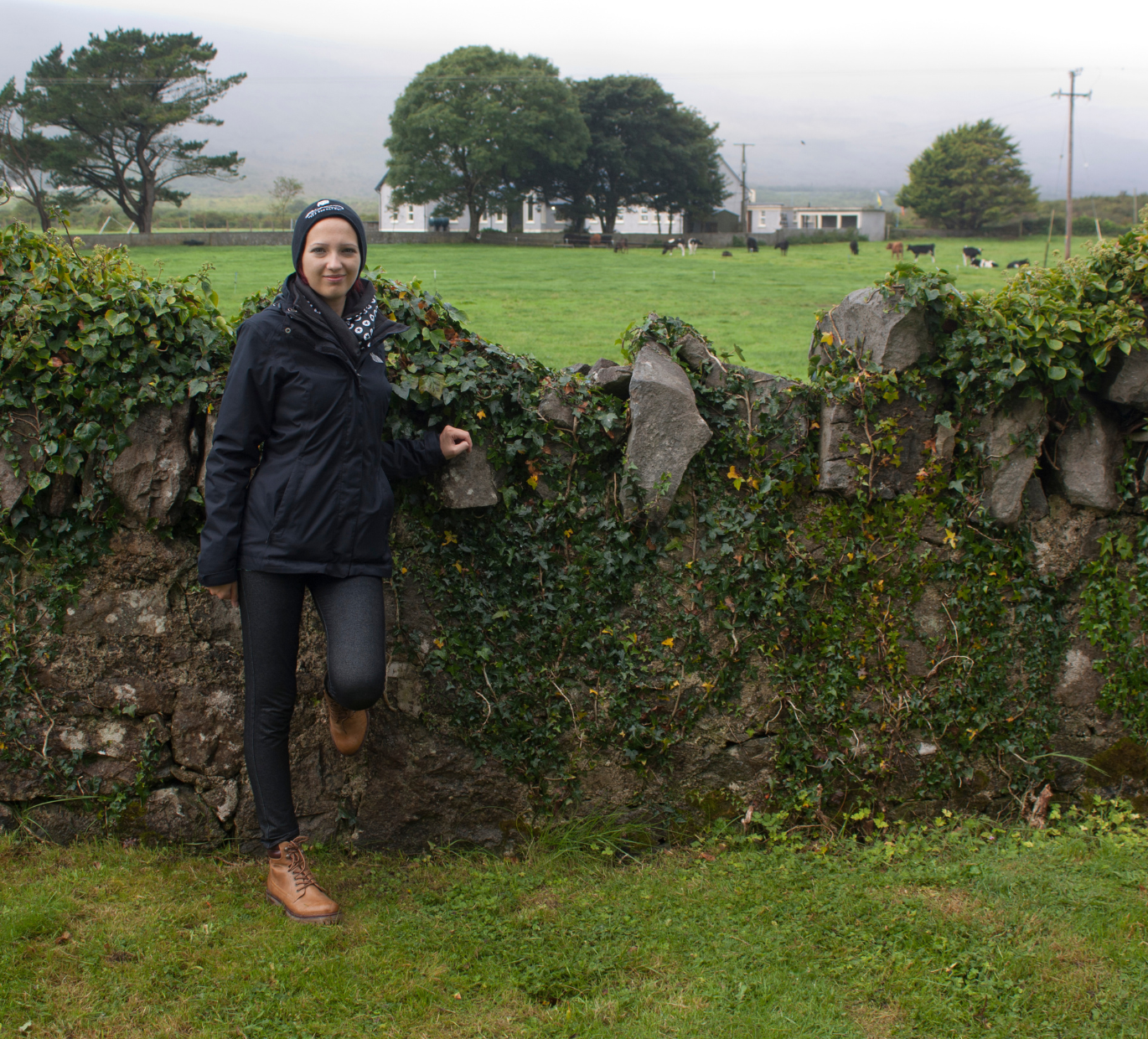 Jessica Jirschik in Irland im Herbst. Sie lehnt an einer alten Steinmauer und trägt Regenjacke und Mütze. Im Hintergrund grasen einige Rinder